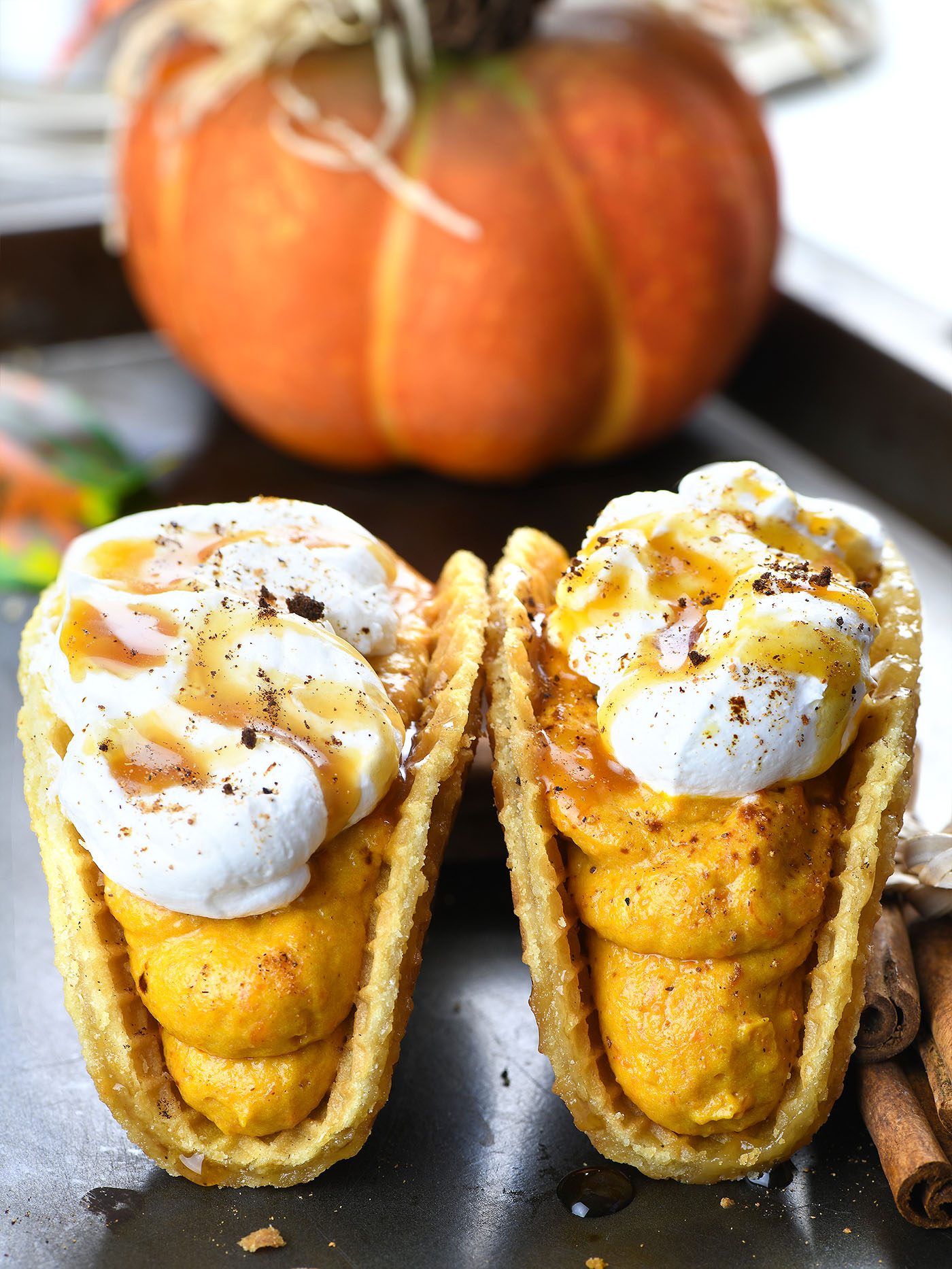 Two pumpkin spice latte tacos with stroopwafel shells filled with creamy pumpkin mousse, topped with whipped cream, caramel drizzle, and a dusting of cinnamon. A pumpkin is visible in the background, adding to the autumnal feel.