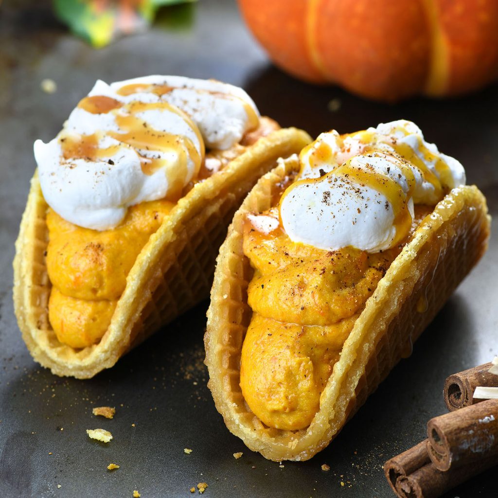 Two pumpkin spice latte tacos with stroopwafel shells filled with creamy pumpkin mousse, topped with whipped cream, a drizzle of caramel sauce, and a sprinkle of cinnamon. A pumpkin and cinnamon sticks are visible in the background, enhancing the fall dessert theme.