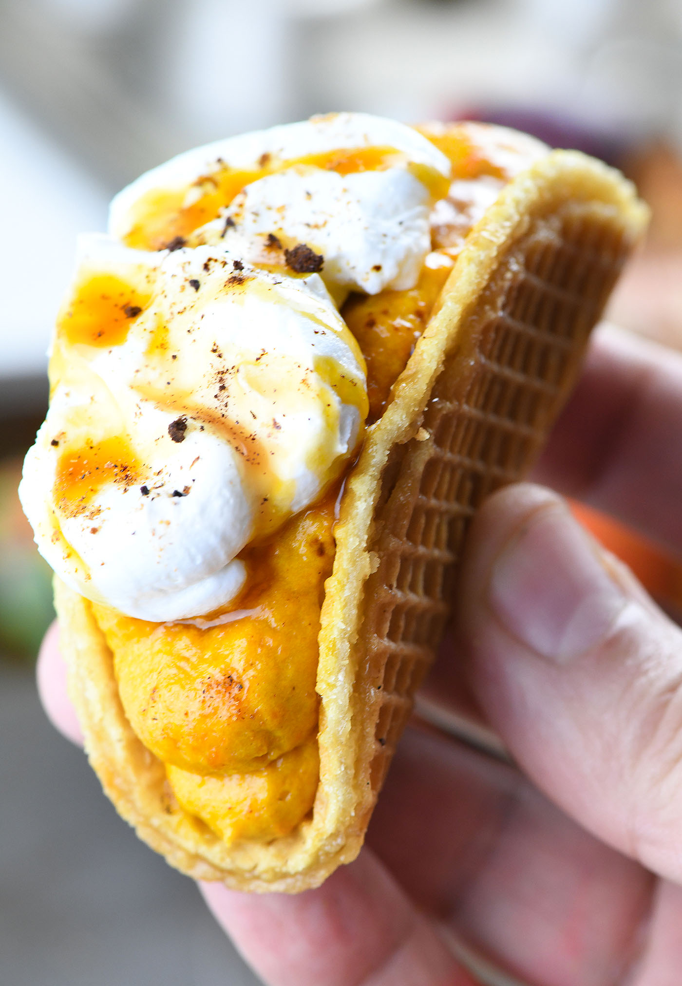 A hand holding a pumpkin spice latte taco with a stroopwafel shell filled with pumpkin mousse, topped with whipped cream, caramel drizzle, and a sprinkle of cinnamon.