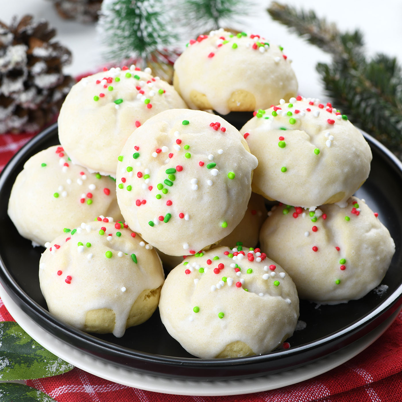 A black plate filled with Italian Christmas cookies, each decorated with white icing and colorful sprinkles in red, green, and white. The festive cookies are set against a holiday-themed background, adding to the Christmas ambiance.