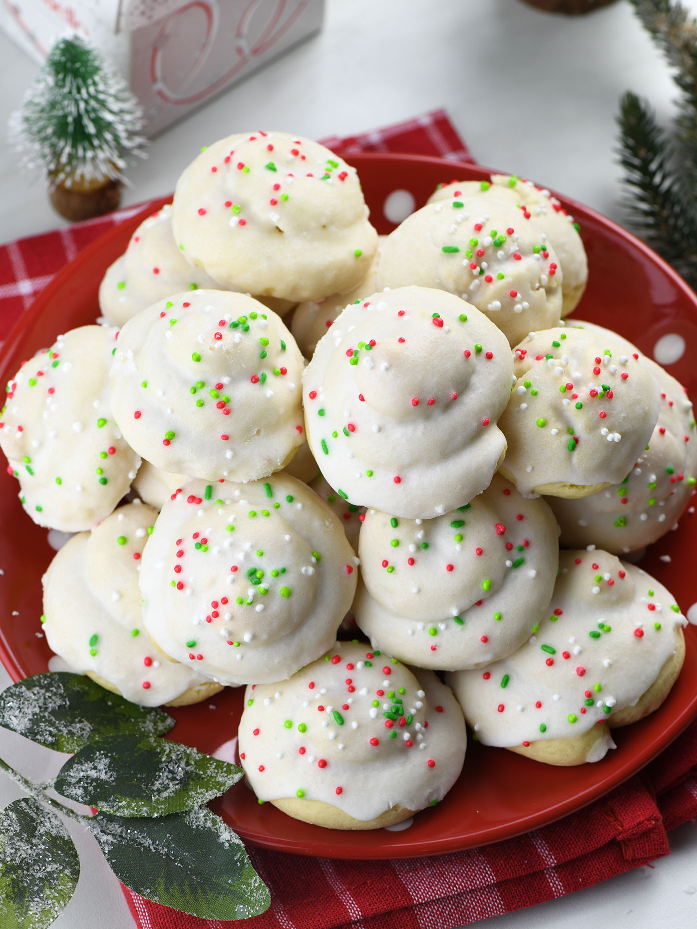 A red plate stacked with Italian Christmas cookies, each topped with white icing and festive red, green, and white sprinkles. The plate is set on a red and white checkered cloth, surrounded by holiday decorations, creating a cozy Christmas atmosphere.