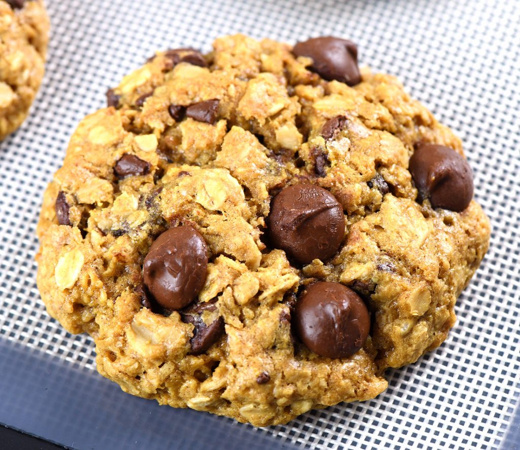 A close-up of a single oatmeal chocolate chip cookie, highlighting its golden brown texture, hearty oats, and gooey chocolate chips on a baking mat.