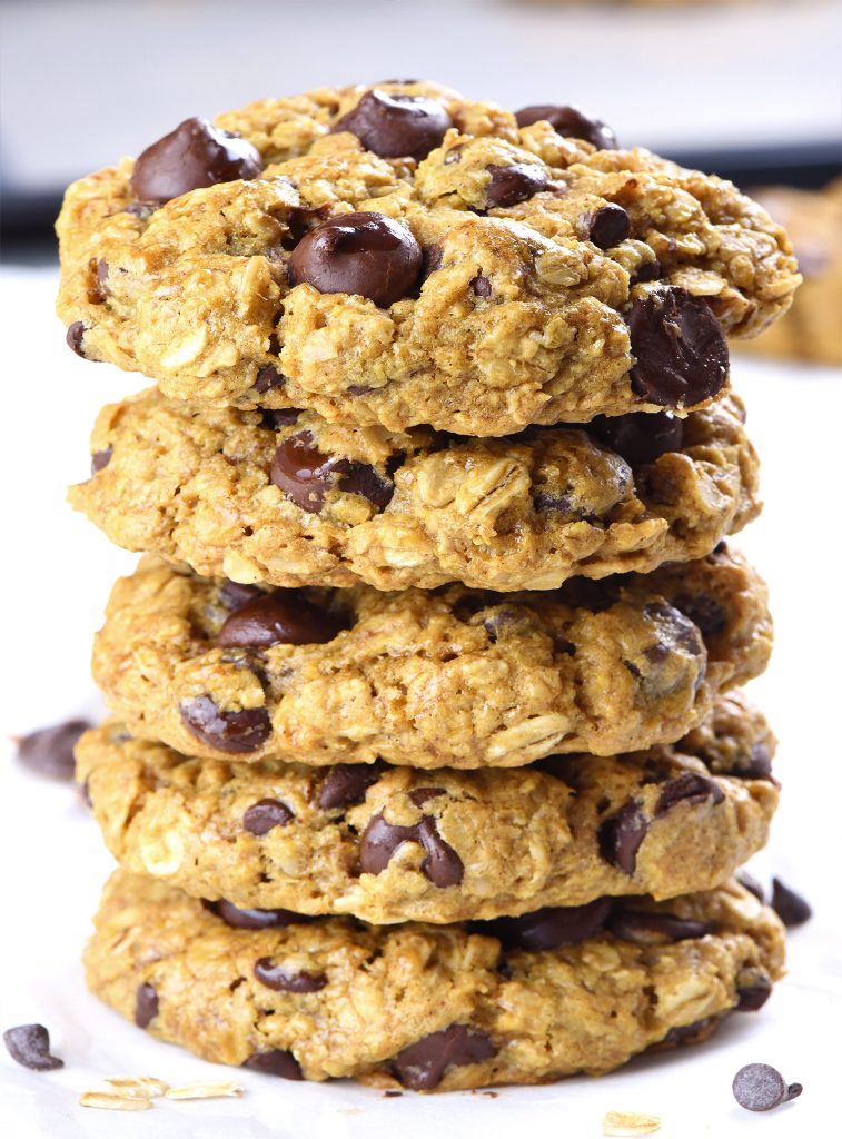 A stack of thick and chewy oatmeal chocolate chip cookies with a golden brown texture and melted chocolate chips, presented on a white background with scattered oats and chocolate chips nearby.