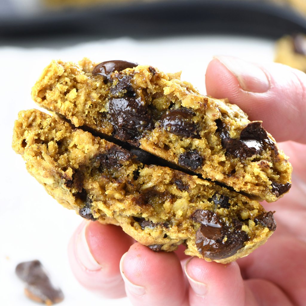 A close-up of a hand breaking open a chewy oatmeal chocolate chip cookie, revealing its gooey chocolate chips and soft, moist interior.