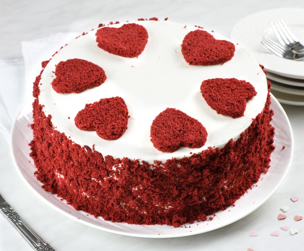 A round red velvet cake with smooth white cream cheese frosting, topped with red heart-shaped cutouts and coated with red velvet crumbs around the sides.
