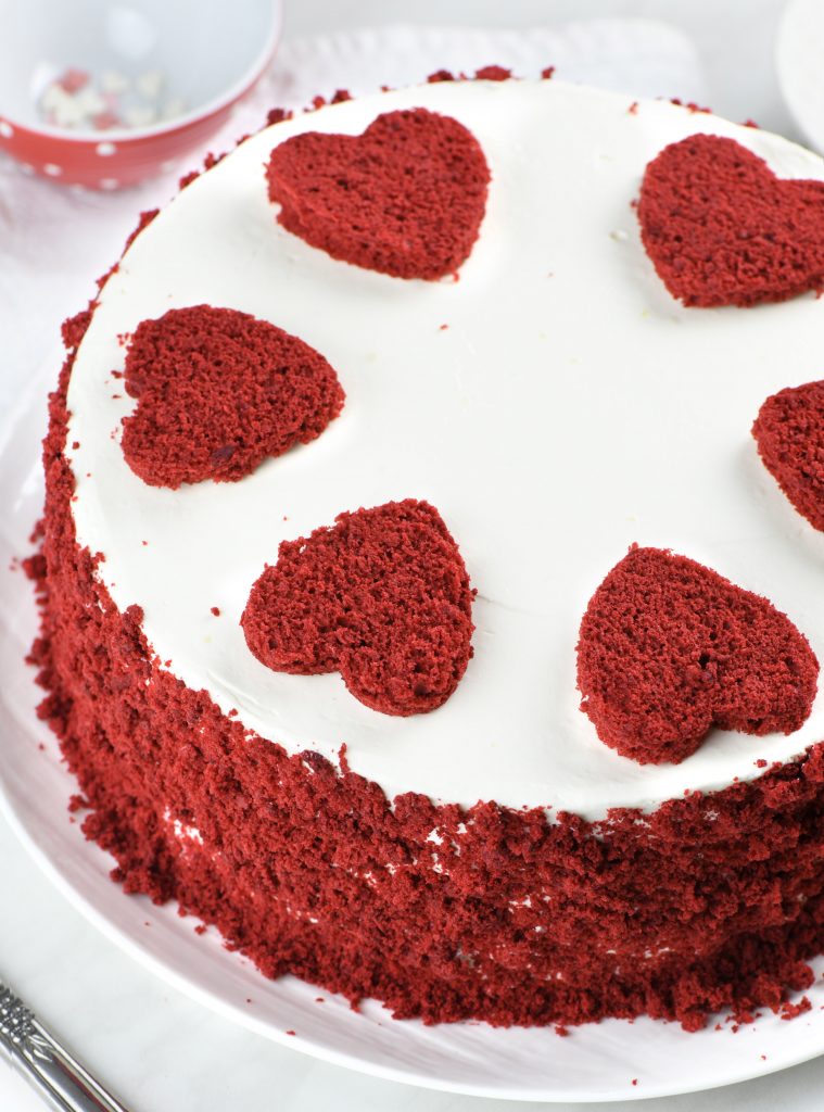 A fully decorated Valentine's Day Cake with white frosting, red velvet crumbs coating the sides, and heart-shaped red velvet cutouts on top.