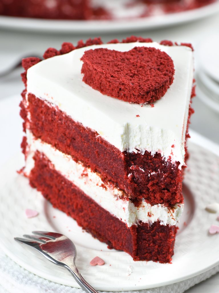 A slice of red velvet cake with creamy white frosting, decorated with a heart-shaped cutout on top, sitting on a white plate with a silver fork beside it.