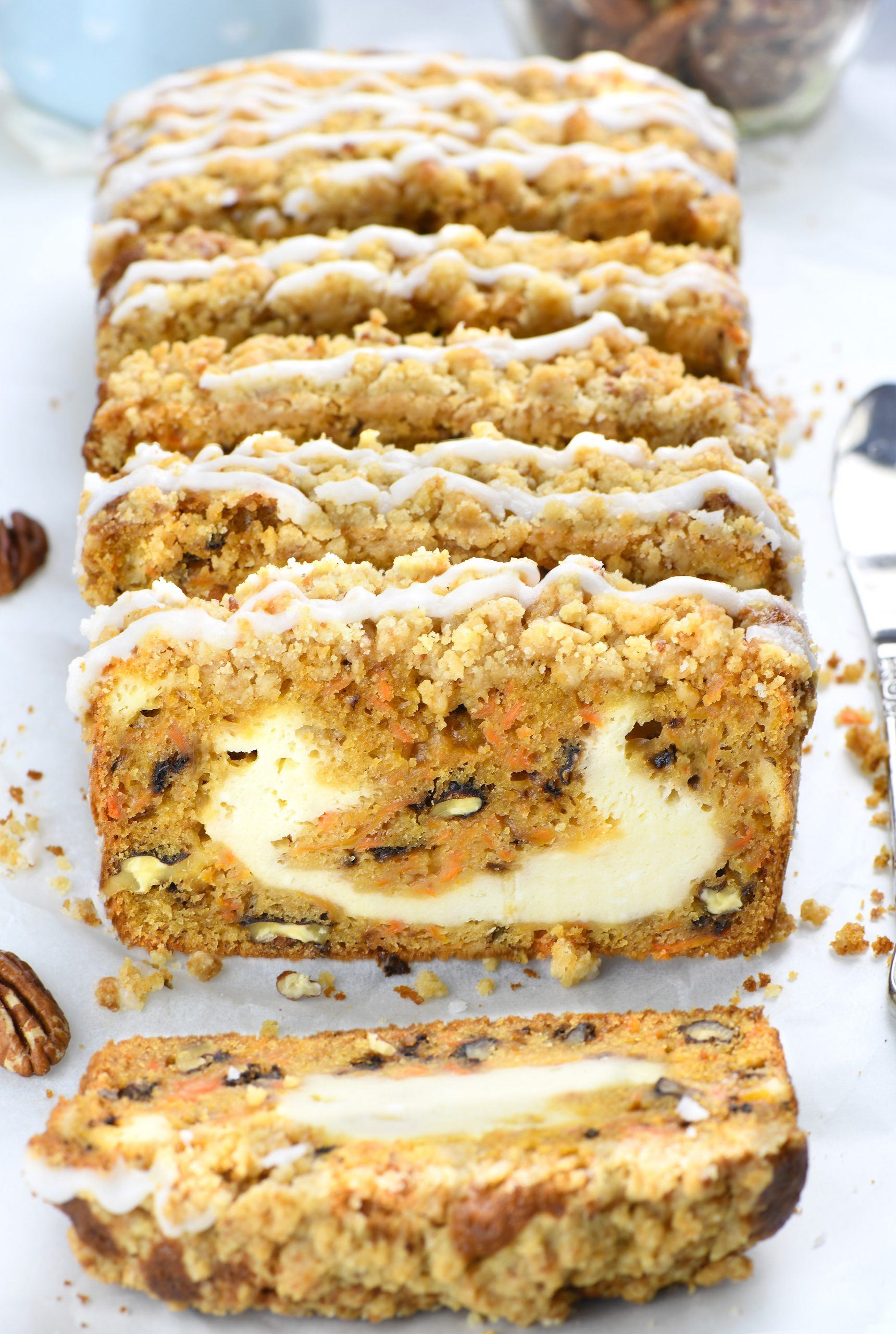 Sliced carrot cake loaf with a creamy cheesecake filling, topped with a crumbly streusel and drizzled with icing. The golden-brown cake contains visible shredded carrots, nuts, and raisins, resting on a parchment-lined surface with pecans scattered around.