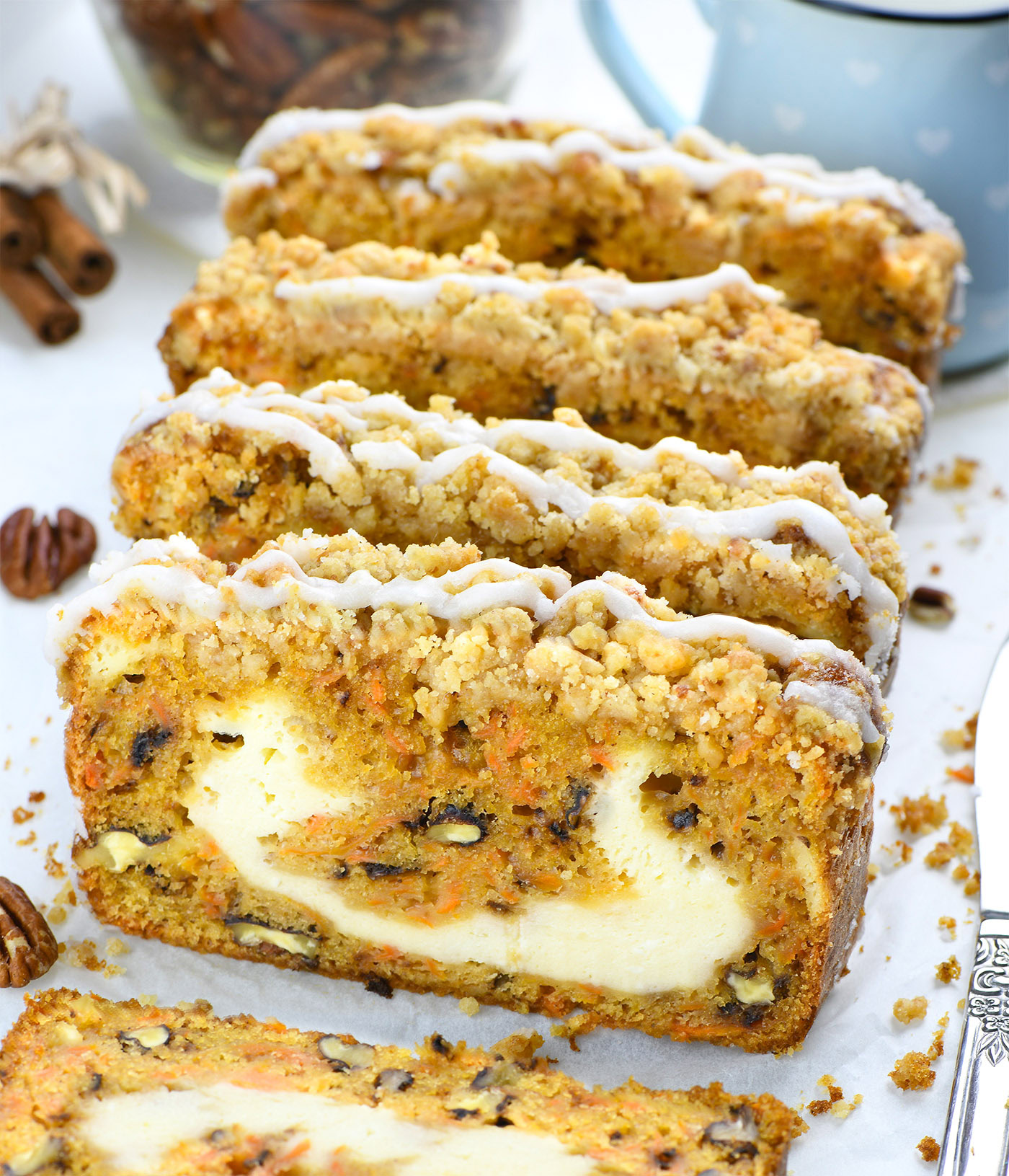 Close-up of a sliced carrot cake loaf with a creamy cheesecake filling, topped with a crumbly streusel and drizzled with icing. The golden-brown cake is packed with shredded carrots, nuts, and raisins. A blue mug with heart patterns and a jar of pecans sit in the blurred background, along with cinnamon sticks.