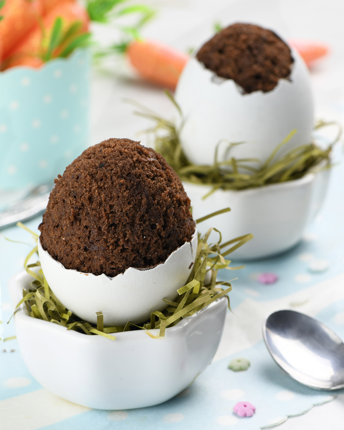 Two chocolate brownies baked inside cracked eggshells, placed on a bed of decorative grass in small white bowls, creating a festive Easter dessert presentation. A pastel background with tiny decorations and faux carrots enhances the holiday theme.