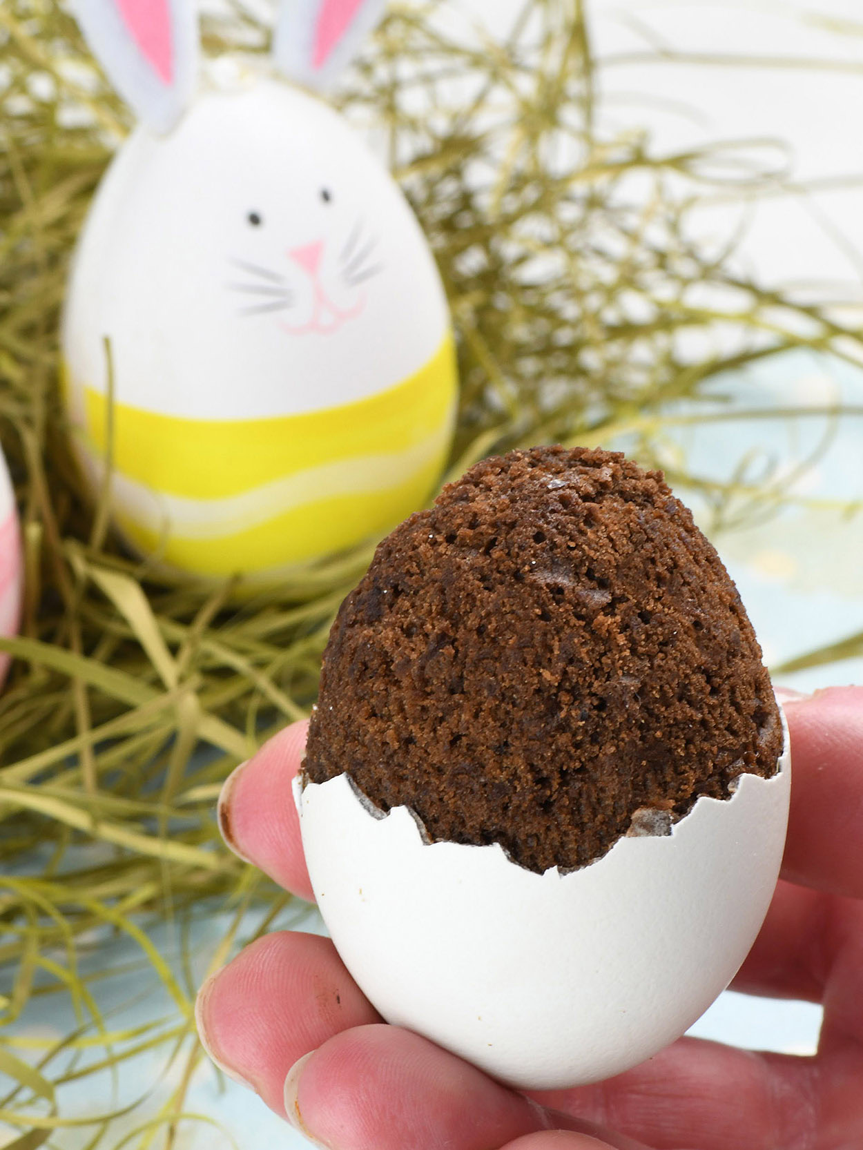 A hand holding a creative Easter dessert—a brownie baked inside a cracked eggshell. The brownie slightly overflows from the top, mimicking the look of a hatching treat. In the background, decorated Easter eggs with bunny faces and pastel colors rest on a bed of hay
