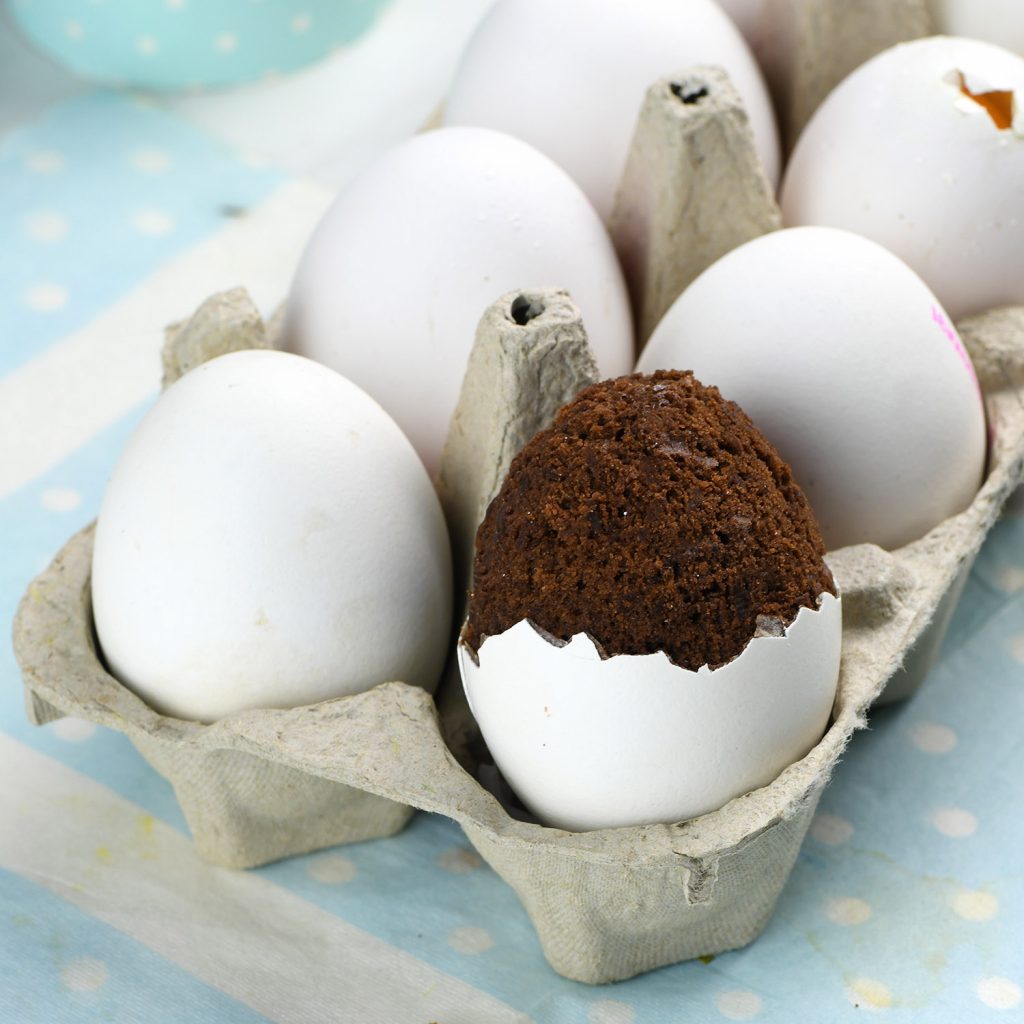 A carton of white eggs with one unique egg featuring a baked chocolate brownie inside, slightly overflowing from the cracked eggshell. The background has a pastel blue polka dot pattern.