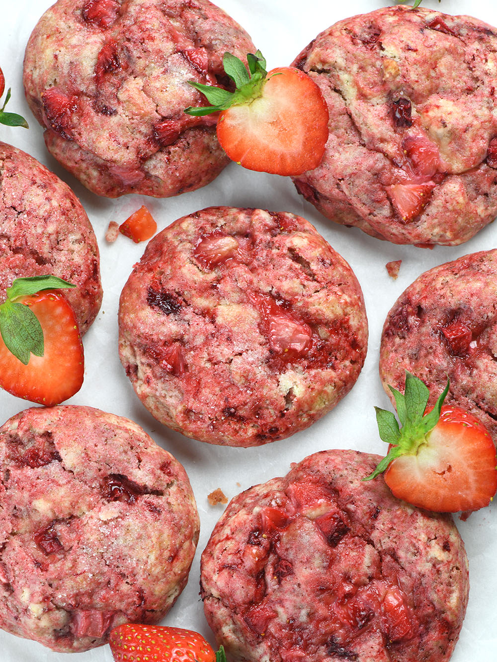 A top-down view of freshly baked strawberry cheesecake cookies with a vibrant pink hue, studded with juicy strawberry pieces, and lightly dusted with sugar. Fresh strawberry slices are scattered around.
