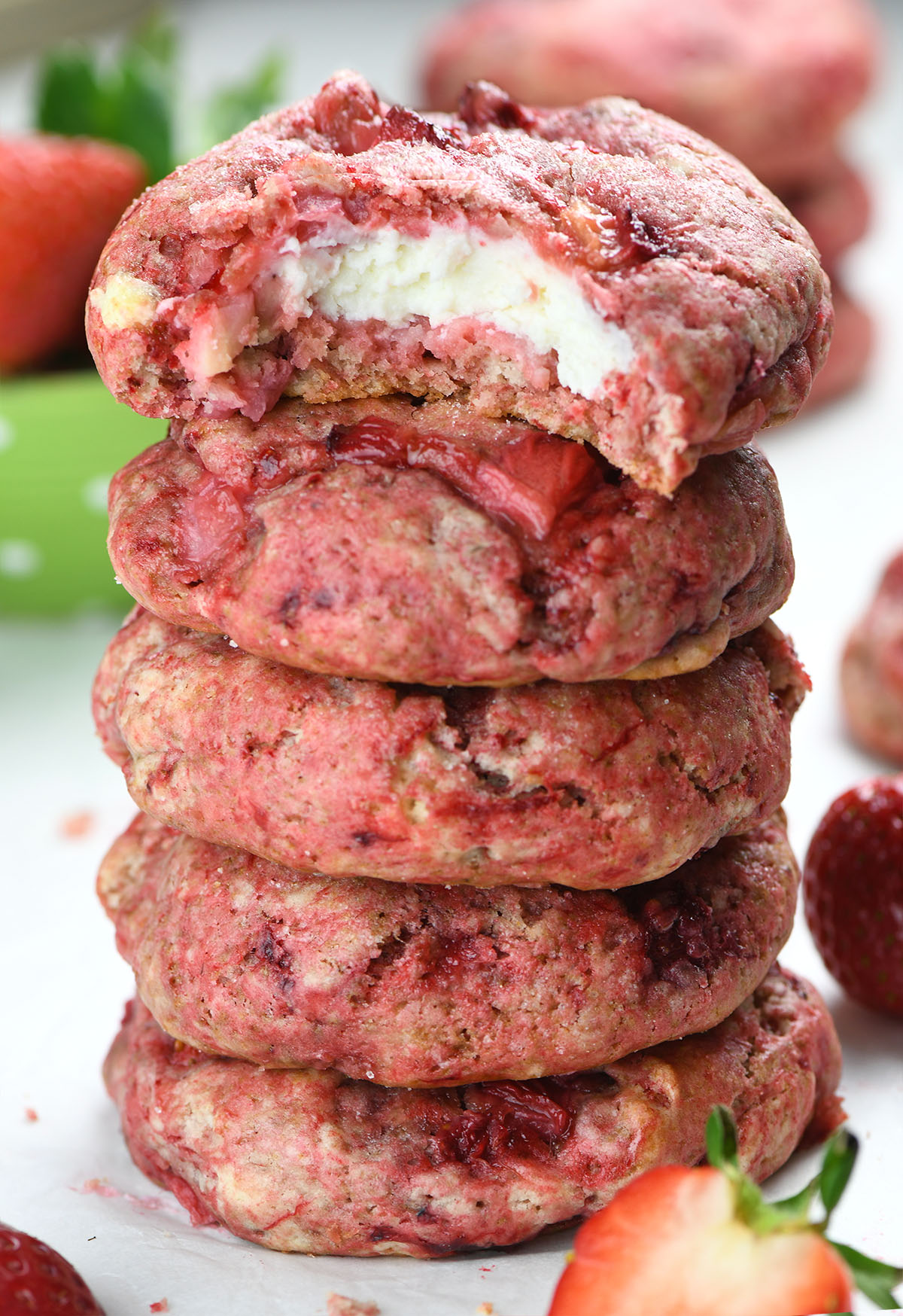 A tall stack of strawberry cheesecake cookies with a bite taken out of the top one, revealing a creamy cheesecake filling. The cookies are vibrant pink with chunks of fresh strawberries, surrounded by whole and sliced strawberries.