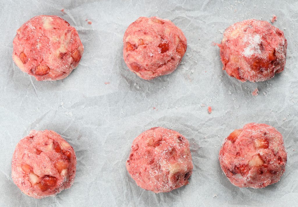 Six balls of strawberry cheesecake cookie dough coated in sugar, arranged on a parchment-lined baking sheet, ready to be baked.
