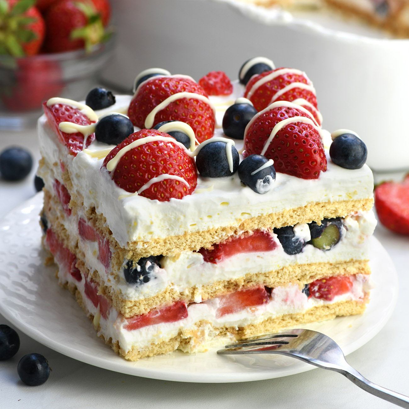 Square image of a layered no-bake berry icebox cake with fresh strawberries and blueberries, whipped cream, and graham crackers, topped with white chocolate drizzle.