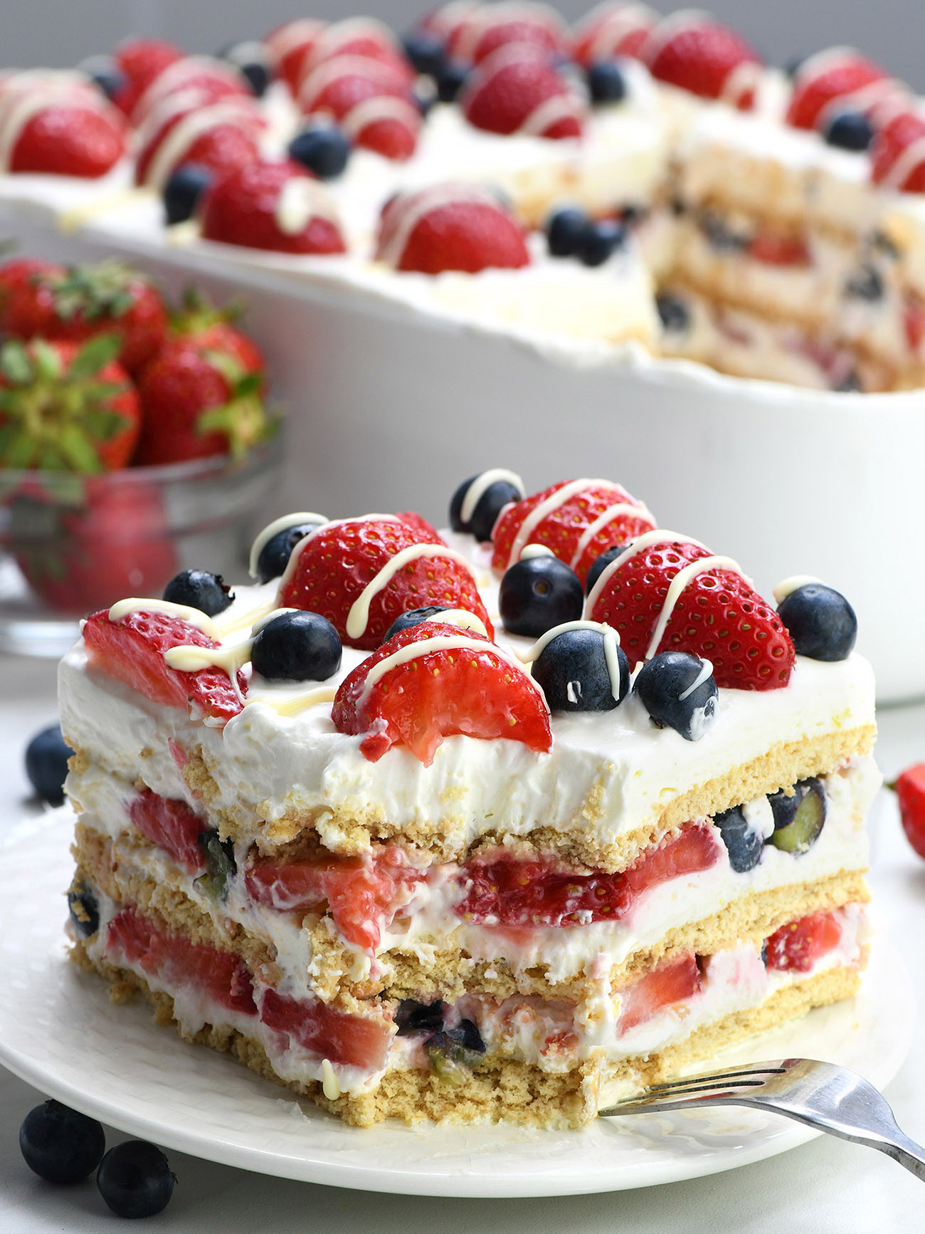Vertical slice of no-bake berry icebox cake with layers of whipped cream, strawberries, blueberries, and graham crackers, topped with fresh fruit and white chocolate drizzle.