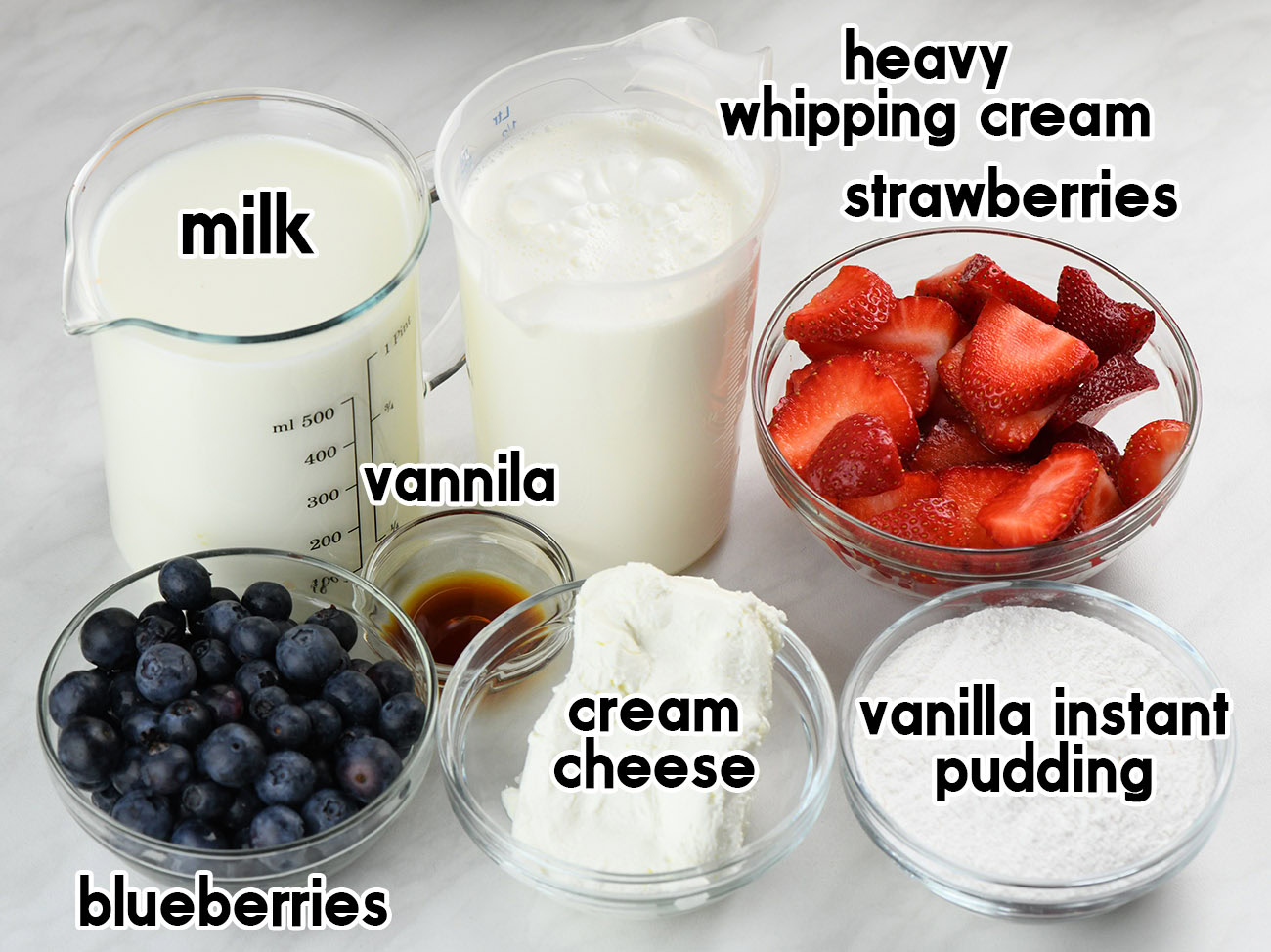 Ingredients for no-bake berry icebox cake arranged on a white surface, including milk, heavy whipping cream, strawberries, blueberries, cream cheese, vanilla extract, and vanilla instant pudding mix.