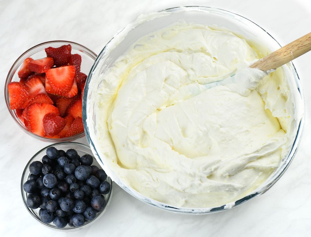 A large bowl of whipped vanilla cream filling with a spatula, next to bowls of sliced strawberries and fresh blueberries, ready for assembling no-bake berry icebox cake.