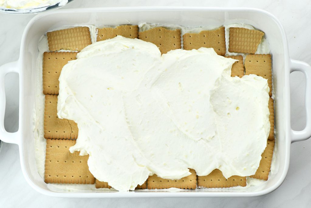 A white casserole dish with a layer of graham crackers topped with whipped vanilla filling, showing the first step in assembling no-bake berry icebox cake.