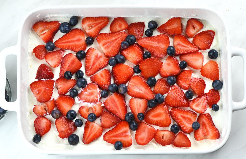Layer of sliced strawberries and blueberries arranged over whipped filling in a white baking dish as part of assembling a no-bake berry icebox cake.
