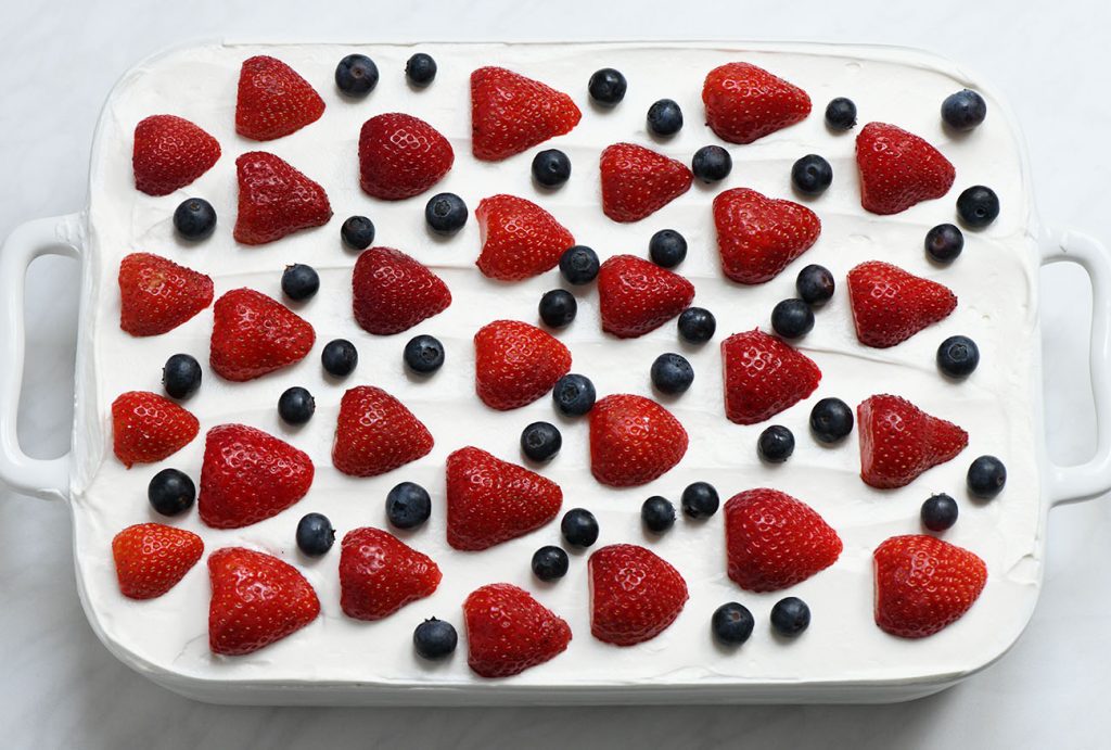 Top view of finished no-bake berry icebox cake in a white dish, topped with halved strawberries and whole blueberries arranged over a smooth whipped cream layer.