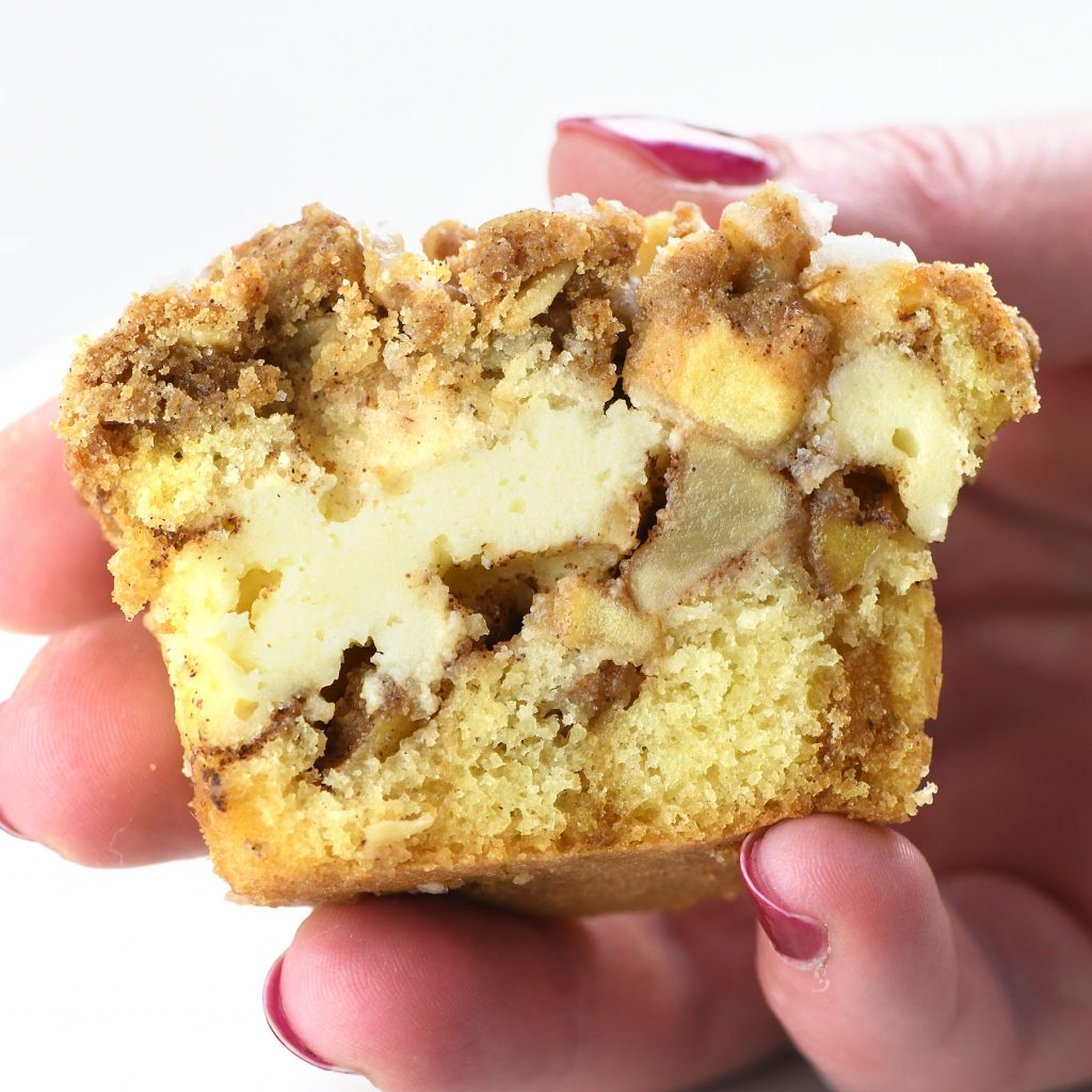 Hand holding half of an apple crisp cheesecake muffin showing layers of moist cake, creamy cheesecake filling, spiced apple chunks, and a crumbly cinnamon streusel topping.