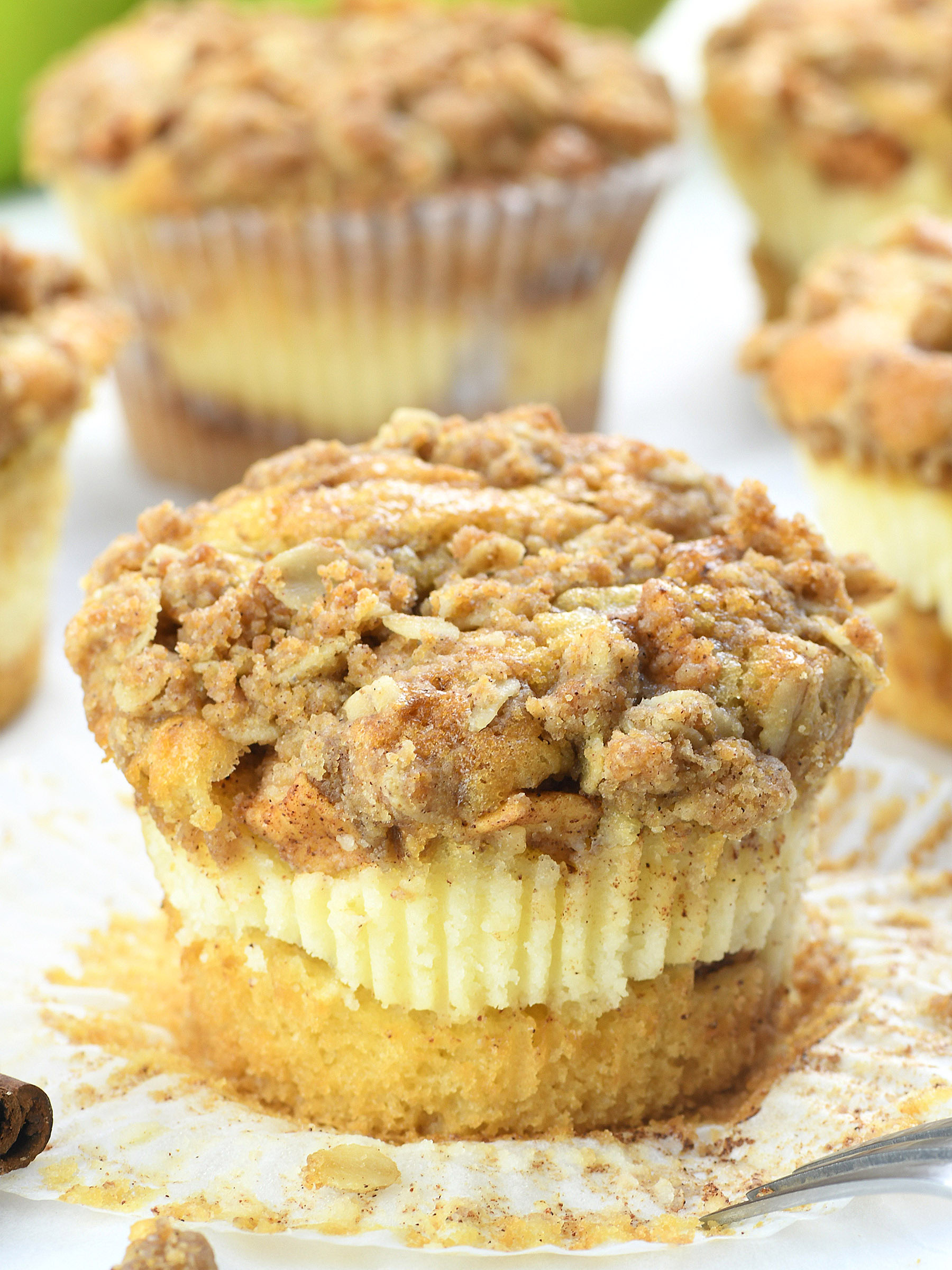 Apple crisp cheesecake muffin with a golden oat streusel topping and creamy cheesecake layer, displayed in a white cupcake liner with cinnamon sprinkles.