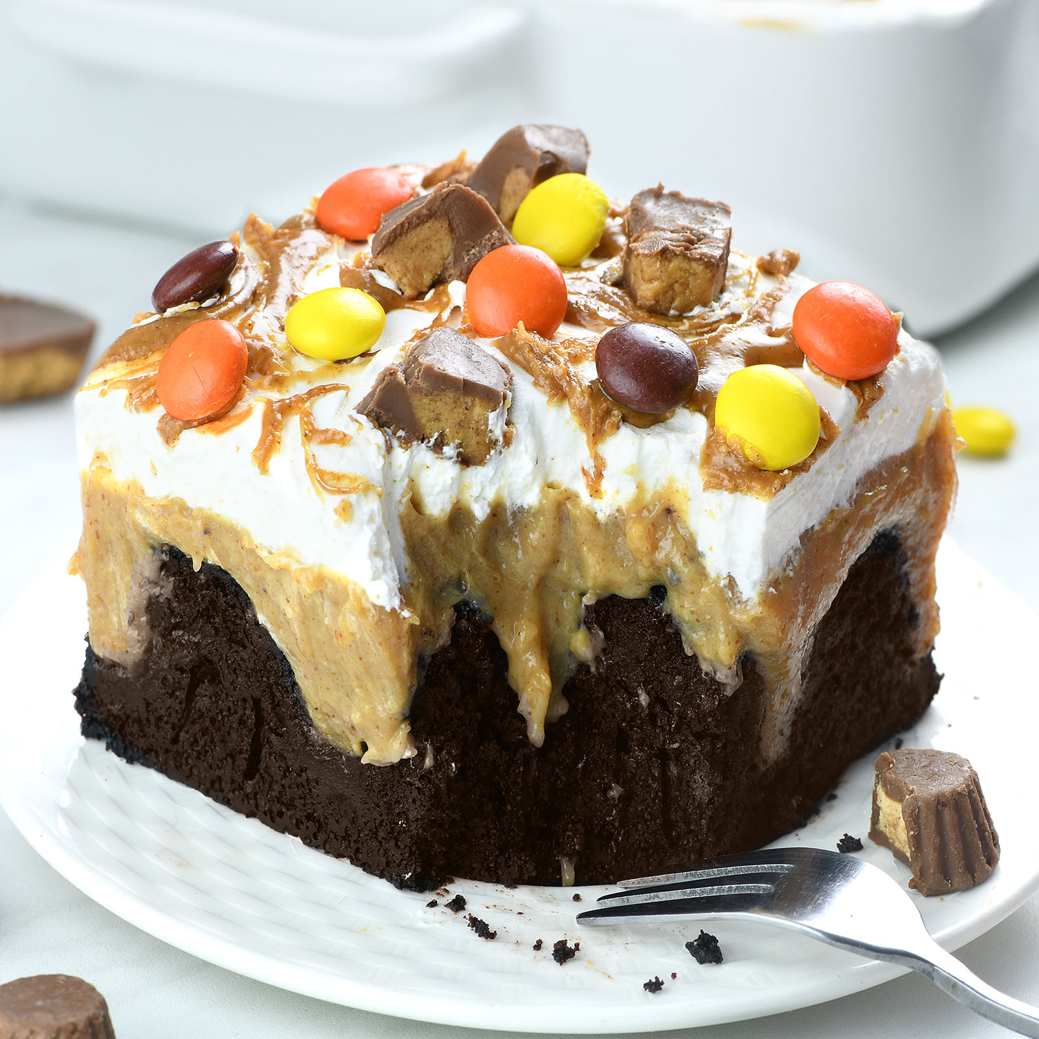 A close-up slice of chocolate peanut butter poke cake on a white plate. The moist chocolate cake is filled with creamy peanut butter filling, topped with fluffy whipped cream, drizzled with peanut butter, and garnished with chopped mini peanut butter cups and colorful Reese’s Pieces