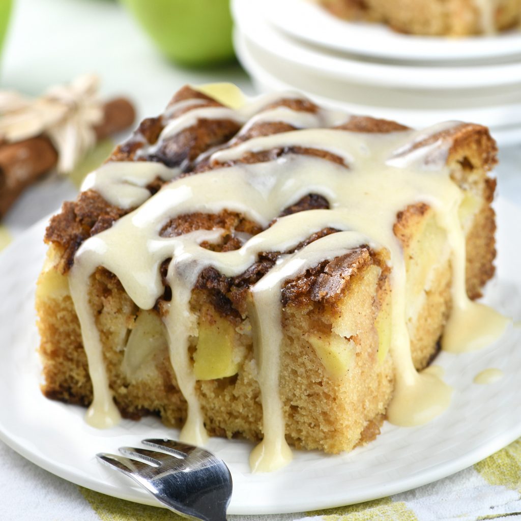 Slice of moist apple cider cake with apple chunks, topped with creamy apple cider glaze, served on a white plate with a fork.