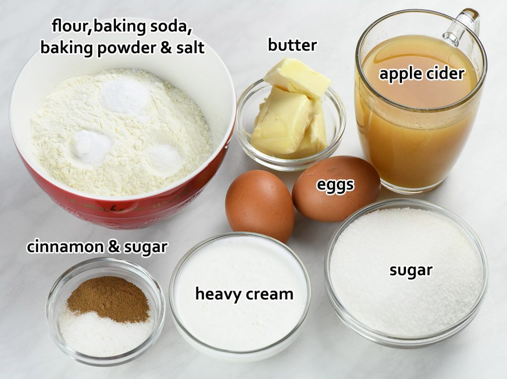 Apple cider cake ingredients displayed on a white surface, including flour with leavening agents, butter, eggs, sugar, heavy cream, cinnamon, and apple cider.