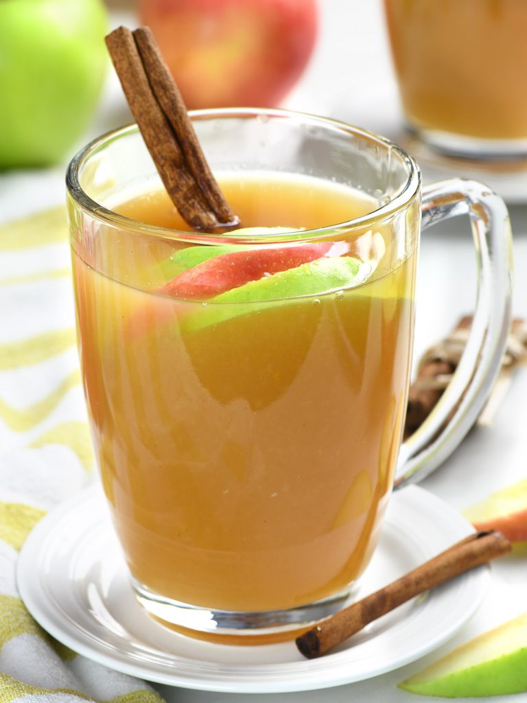 Glass mug of easy homemade apple cider garnished with fresh apple slices and a cinnamon stick, served on a white saucer with extra apple slices and cinnamon nearby.