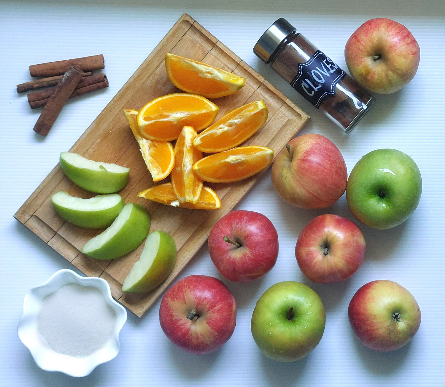 Fresh ingredients for homemade apple cider, including whole red and green apples, orange slices on a cutting board, cinnamon sticks, ground cloves, and sugar.