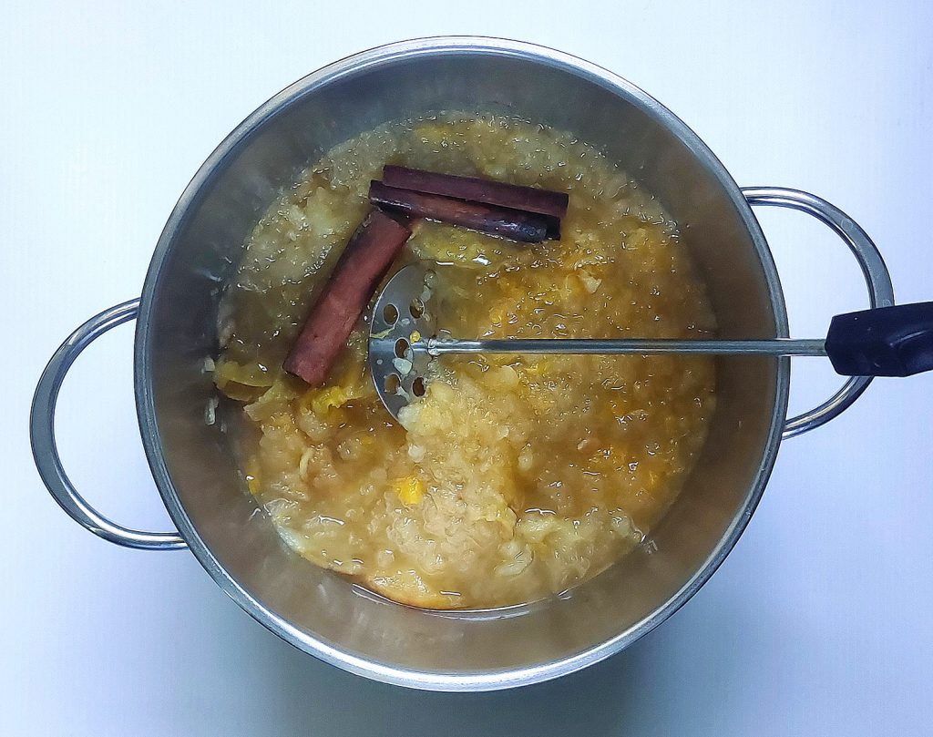 Mashing simmered apples, oranges, and cinnamon sticks in a pot to make homemade apple cider.