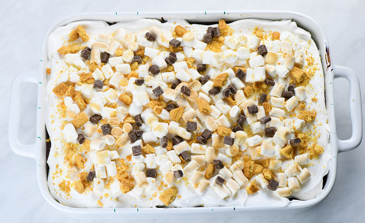 A pan of S’mores poke cake topped with fluffy whipped cream, mini marshmallows, graham cracker pieces, and chocolate chunks.