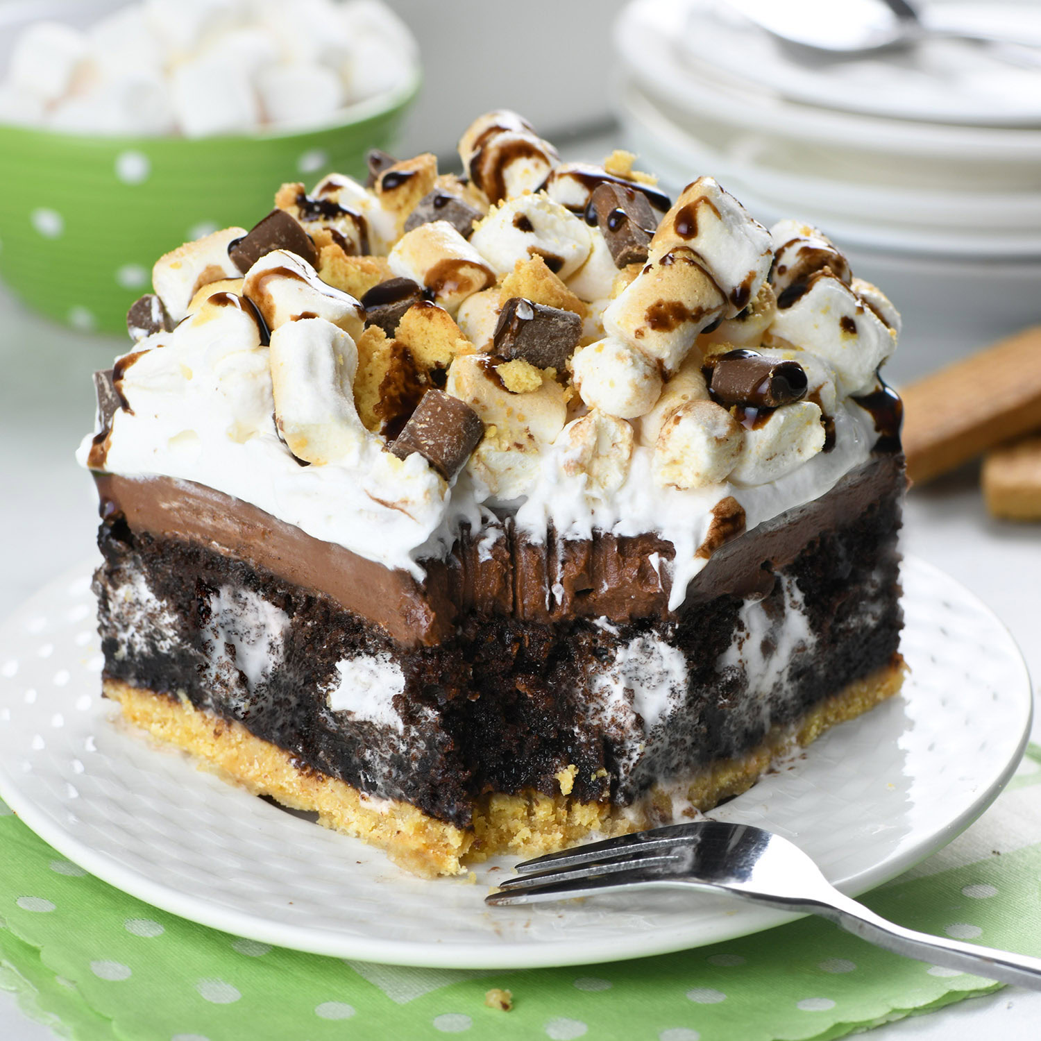 A delicious slice of S’mores poke cake with layers of chocolate cake, gooey marshmallow filling, and a graham cracker crust, topped with whipped cream, mini marshmallows, and a drizzle of chocolate.